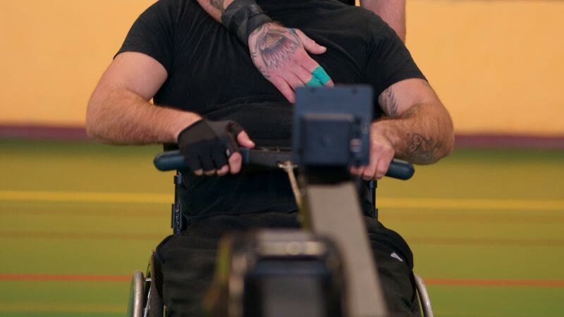 Un adhérent en fauteuil tire la poignée d'un rameur, la main de son binôme posée sur son épaule.