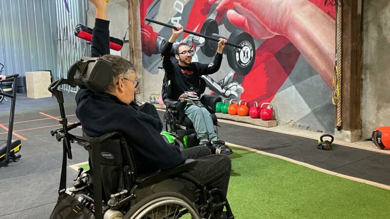 Un adhérent en fauteuil roulant pousse une barre au-dessus de sa tête, encouragé par un autre adhérent.