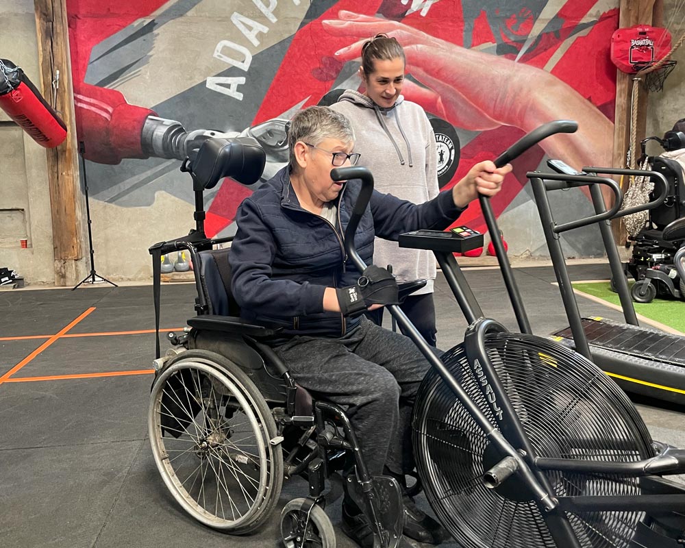 Une coach accompagne une adhérente en fauteuil roulant sur un vélo à air, pendant une séance dans la salle d'ADAPTATEAM.