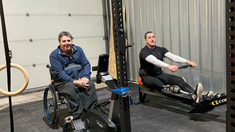 Deux adhérents côte à côte dans la salle, l'un en fauteuil sur un vélo, l'autre équipé d'une prothèse de jambe sur un rameur, souriants.